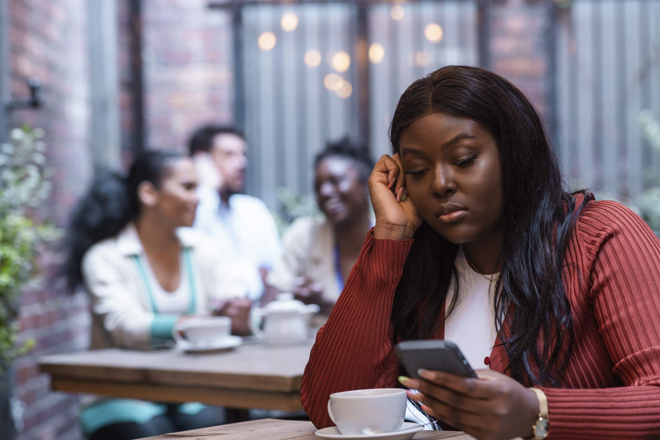 Young African-American woman on phone sitting at a table alone, with long black hair, white t-shirt, and red cardigan.