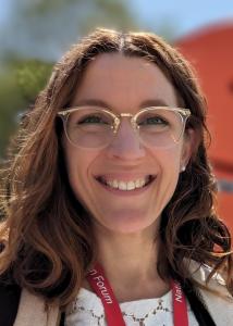 Anne Pisor headshot with long wavy brown hair and gold glasses, smiling.