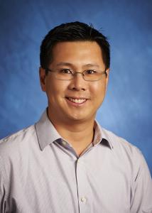 Headshot of Carlomagno Panlilio in front of blue backdrop with glasses and short dark hair wearing a light purple dress shirt. 