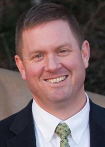 Headshot of Daniel Perkins with short brown hair, a white shirt, green and blue patterned tie, and a black jacket.