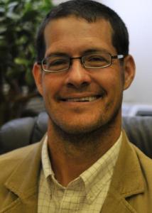 Headshot of Frank Hillary with black hair, glasses, white and tan checkered shirt, and tan jacket.