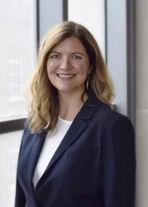 Emily Ansell head shot in navy suit in front of window.