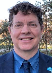 Headshot of Orfeu Buxton outside in thin-rimmed, light-colored glasses wearing a blue shirt, patterned tie and dark jacket.