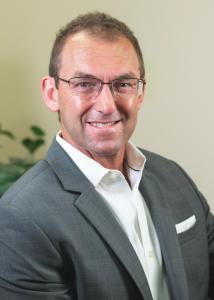 Headshot of Paul Riccomini, a white man with short hair, glasses, and a grey jacket with white shirt.