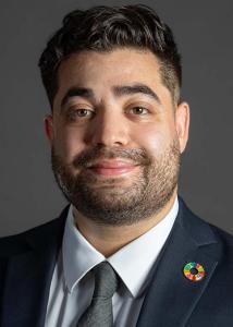 Headshot of Mark Ortiz, who has short, dark hair, a beard, and is wearing a blue suit jacket with a white collared shirt and grey tie.